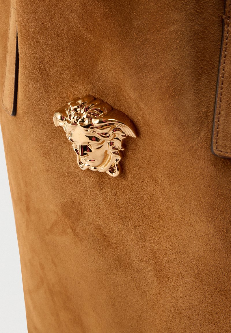 Braune Wildlederhandtasche mit einem auffälligen goldenen Medusa-Kopf als Verschluss, mit weicher Textur und minimalistischem Design.