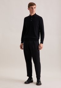 Schwarzer Pullover mit Kragen und drei Knöpfen, kombiniert mit locker sitzenden dunklen Hosen und schwarzen Lederschuhen. Glatte Textur.