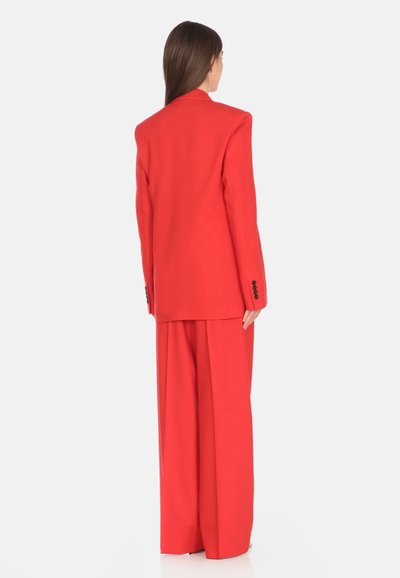 Completo su misura rosso con giacca strutturata, spalline e pantaloni a gamba larga, realizzato in tessuto morbido, che mette in risalto una silhouette pulita.