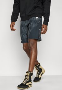 Μαύρο αθλητικό σορτς με κυματιστό μοτίβο σε σκούρο γκρι, με διακριτική λευκή λεπτομέρεια. Συνδυασμένο με μαύρα και μπεζ ψηλά παπούτσια.