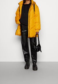 Abrigo acolchado de color amarillo brillante con capucha, combinado con una sudadera negra, pantalones de cuero negros y zapatos negros. Lleva un pequeño bolso negro.