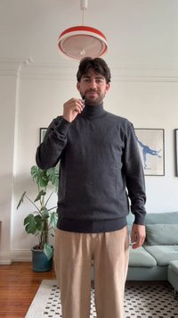 Gray turtleneck sweater, ribbed cuffs and hem, paired with beige corduroy pants. Standing indoors with a plant and couch in the background.