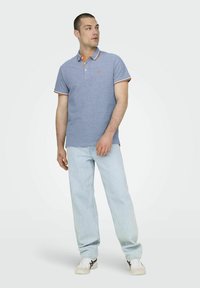 Camisa polo azul claro con acentos naranjas, de manga corta y un logotipo bordado sutil. Combinada con jeans de mezclilla claros y zapatillas blancas.