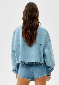 Chaqueta de mezclilla azul claro, corta y con hombros caídos, con el dobladillo deshilachado, combinada con pantalones cortos de cintura alta a juego. La textura parece suave y desgastada.