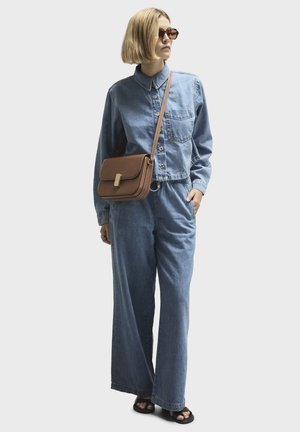 Kvinde iført denimjakke og bukser med vide ben, brun crossbody-taske, solbriller, sorte sandaler, stående med den ene hånd i lommen, ser mod højre.