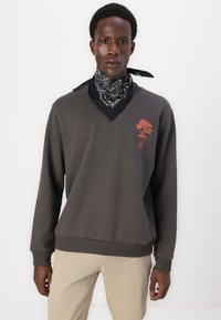 Grå sweatshirt med långa ärmar, design med v-ringning och röd trädgrafik. Kombinerad med en svart bandana med vita mönster.