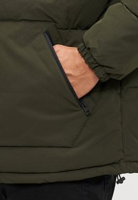 Veste matelassée verte isolée avec une poche zippée et des poignets élastiques ; texture lisse et tissu durable. Coutures accentuées visibles.