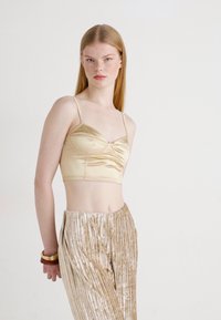Satin kratki top u zlatnoj boji s naboranim prednjim detaljem i tankim trakama, u kombinaciji sa zlatnim plišanim hlačama s nabranim detaljima. Glatka tekstura i elegantan dizajn.