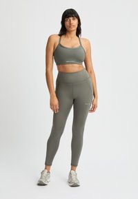 Reggiseno sportivo verde oliva e leggings a vita alta realizzati in tessuto elasticizzato, con un branding minimalista. Il modello indossa scarpe sportive.