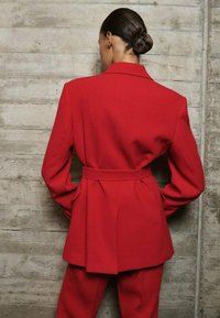 Woman with dark hair in a low bun wears a red belted blazer and matching trousers, standing against a textured concrete wall, facing away.