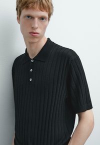 Polo shirt nero a costine realizzato in tessuto a maglia, dotato di colletto, chiusura con tre bottoni e maniche corte con un design aderente.