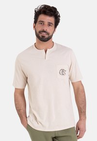 T-shirt beige clair à manches courtes avec un col henley peu profond et une poche poitrine avec un design imprimé. Texture douce, semblable au coton.