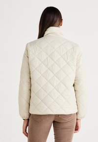 Lysebeige quiltet jakke med høj krave, elastiske manchetter og mønster af diamanter. Har en afslappet pasform og en blød, glat tekstur.