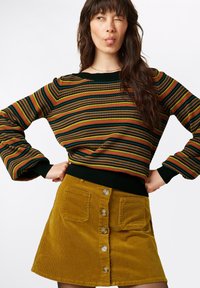 Sweter w paski w zielonych, czerwonych i żółtych odcieniach, zestawiony z musztardową żółtą mini spódnicą z korduroy, z zamknięciem na guziki z przodu i kieszeniami.