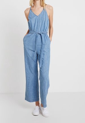 Mujer con mono sin mangas de mezclilla azul claro con escote en V, cinturón en la cintura, pantalones anchos y zapatillas blancas, de pie contra un fondo liso.