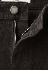 Prim-plan cu pantaloni de catifea côtelată maro închis, cu fermoar metalic parțial deschis și nasture metalic cu marcă.