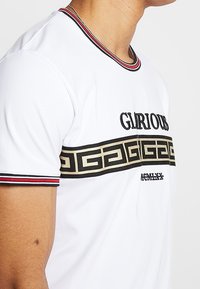 Valkoinen lyhythihainen t-paita, jossa on ribbien punainen ja musta kaulus. T-paidassa on musta ja kultainen graafinen nauha, jossa lukee "MAHTAVA".