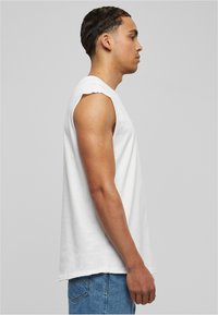 T-shirt en coton blanc sans manches avec des bords bruts, présentant une coupe décontractée. Porté avec un jean en denim bleu, vue de profil sur fond uni.