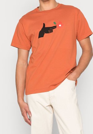 Man draagt een roestoranje t-shirt met een zwarte handafbeelding die naar een kleine rode bloem wijst, gecombineerd met ecru broek.