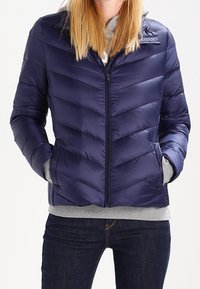 Tmavě modrá prošívaná bunda s chevronovým vzorem šití, zapínáním na zip a kapsami. Nasažená přes šedou mikinu s kapucí. Měkký, lesklý nylonový materiál.