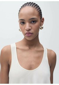 A model wears a light beige tank top with a wide neckline and thin straps, accentuated by large gold hoop earrings.