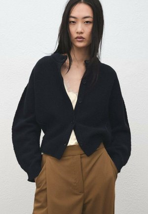 Cardigan negro de textura suave, con diseño sin cuello y botones delanteros, llevado sobre una camiseta crema y pantalones marrones.