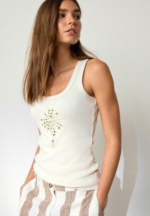 Hosszú barna hajú nő, fehér tank topot visel, melyen tükröződő virágminta van, és bézs-fehér csíkos nadrágot, kezét a zsebében.