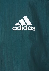 Logo Adidas blanc avec trois bandes diagonales et texte sur fond de tissu texturé bleu sarcelle.