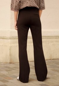 Schwarze Schlaghosen mit einer glatten Textur, ausgestattet mit einer Gesäßtasche. Kombiniert mit einem Leopardenmuster-Oberteil und hellen Schuhen auf einem gepflasterten Boden.
