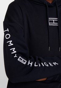 Navi sinine kapuutsiga pusa, millel on tõmblukk, valge tikandiga "TOMMY HILFIGER" tekst varrukal ning logo detail ees.