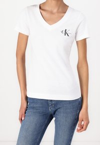 Camiseta de algodón blanca con diseño de cuello en V, mangas cortas y un pequeño logo negro de Calvin Klein Jeans en la parte izquierda del pecho.