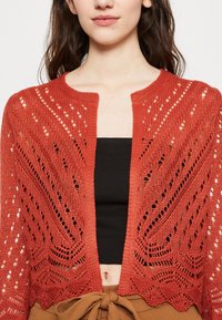 Cardigan rosso lavorato a maglia con design a giorno, dotato di scollo rotondo e maniche a tre quarti. Texture a fantasia con vestibilità ampia.