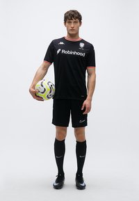 Melnas futbola tērps ar sarkaniem akcentiem, tajā ir Kappa logotips, Nike šorti, melnas zeķes un rokā turēta rakstaina futbola bumba.