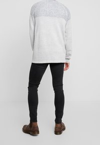 Světlý šedý pletený svetr s texturovanou zadní částí v oblasti ramen, doplněný černými úzkými kalhotami a hnědými kotníkovými botami.