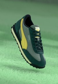 Vihreä ja keltainen Puma-tennari roikkuu vihreän tekstuurin päällä, näyttäen sivuprofiilin, jossa kengän nauhat ja logo ovat näkyvissä.