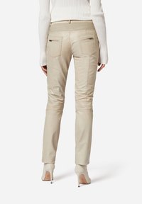 Beige Lederhosen mit Slim Fit, strukturiertem Rücken, zwei Reißverschlusstaschen und elastischem Bund, kombiniert mit weißen High Heels.