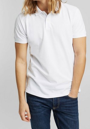 Man draagt een effen wit poloshirt met korte mouwen en donkerblauwe spijkerbroek, staand met één hand in zijn broekzak tegen een grijze achtergrond.