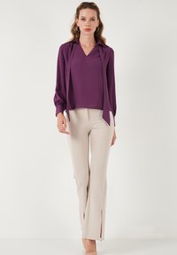 Blusa morada con escote en V y mangas fluidas, combinada con pantalones beige de campana con aberturas laterales en el dobladillo.