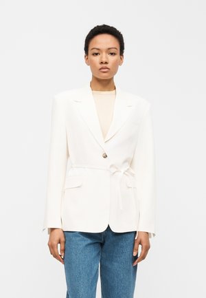 Young person wearing a white blazer with a belt tied at the waist and blue jeans, standing against a plain white background.