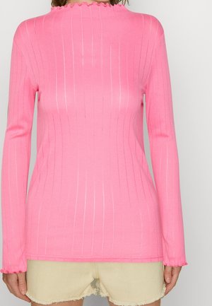 Persoon draagt een nauwsluitend roze ribgebreid shirt met lange mouwen en een gerimpelde rand bij de halslijn en manchetten, gecombineerd met lichtbeige versleten korte broek.