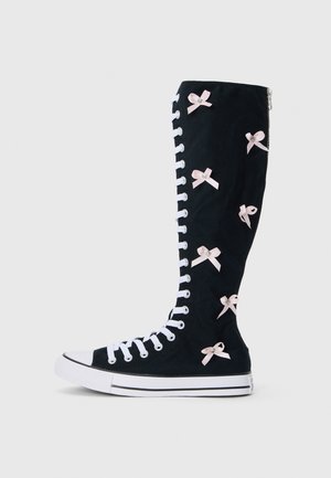 Černé plátěné vysoké boty s bílými šňůrkami, které mají růžové saténové mašle podél holeně a boční zip. Pevná gumová podrážka se šedými akcenty.