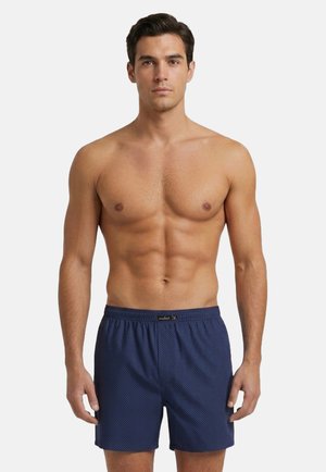 Sportlicher Mann mit kurzen dunklen Haaren steht und trägt dunkelblaue gemusterte Boxershorts vor weißem Hintergrund.