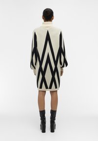 Gebreide jurk met een hoge kraag, voorzien van een crème basis en gedurfde zwarte geometrische patronen. Middenlang met lange, volumineuze mouwen.