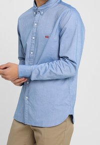 Homme portant une chemise à manches longues bleu clair avec le logo rouge Levi's sur la poitrine et un pantalon beige, ajustant le poignet avec les mains.