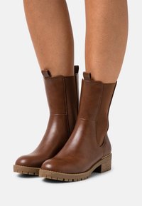 Botines de cuero marrón con paneles laterales elásticos, puntera redondeada, suela texturizada y tacón apilado. La parte superior es lisa con un acabado brillante.