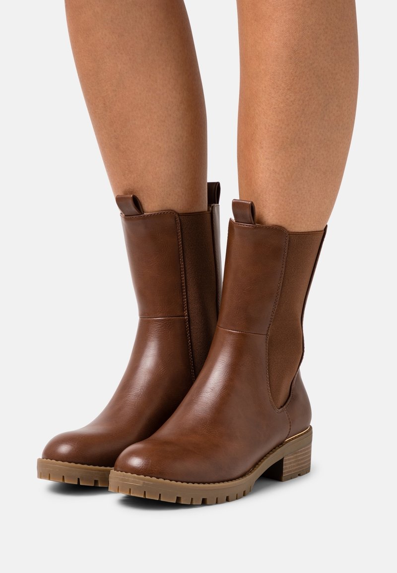 Botines de cuero marrón con paneles laterales elásticos, puntera redondeada, suela texturizada y tacón apilado. La parte superior es lisa con un acabado brillante.