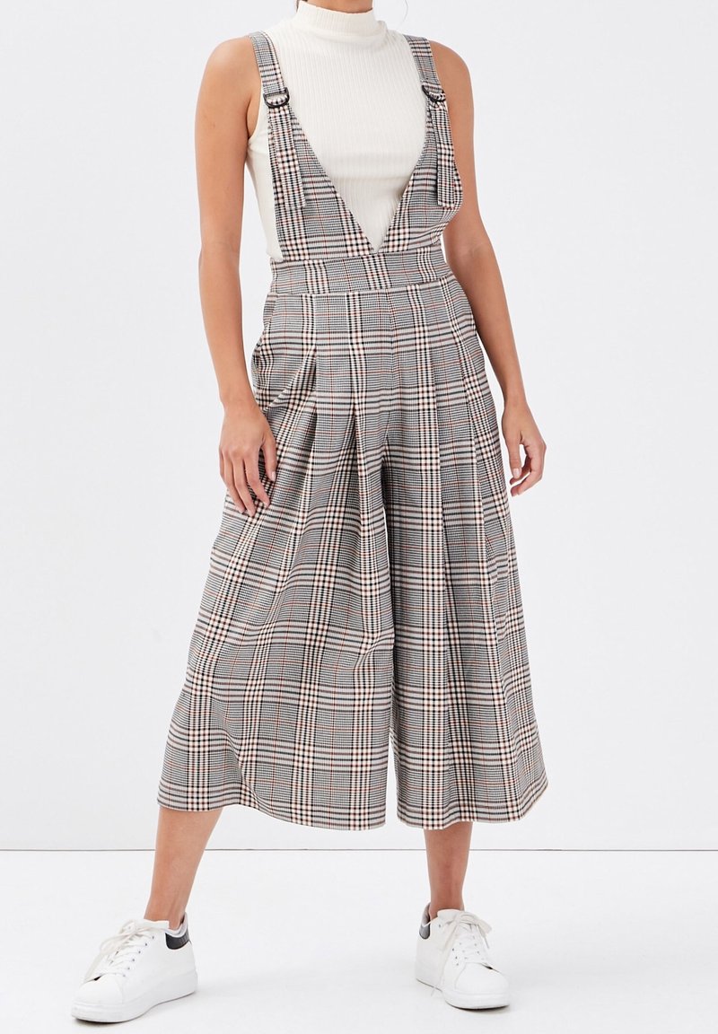 Femme portant un pantalon à bretelles large à carreaux avec un haut blanc sans manches à col roulé et des baskets blanches, debout devant un fond uni.
