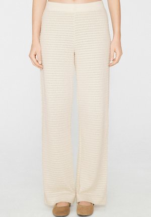 Kremowe szerokie dzianinowe spodnie noszone przez stojącą osobę z rękami swobodnie opuszczonymi wzdłuż boków i brązowymi płaskimi butami.