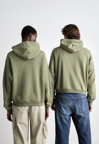 Sudaderas verde oliva con un corte relajado y capuchas con cordón, combinadas con pantalones cargo color beige claro y jeans de mezclilla azules, vistas desde atrás.