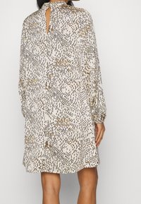 Robe à manches longues en tissu beige clair avec un motif texturé de accents noirs et dorés ; présent une attache au cou au dos et un détail en forme de trou de serrure.